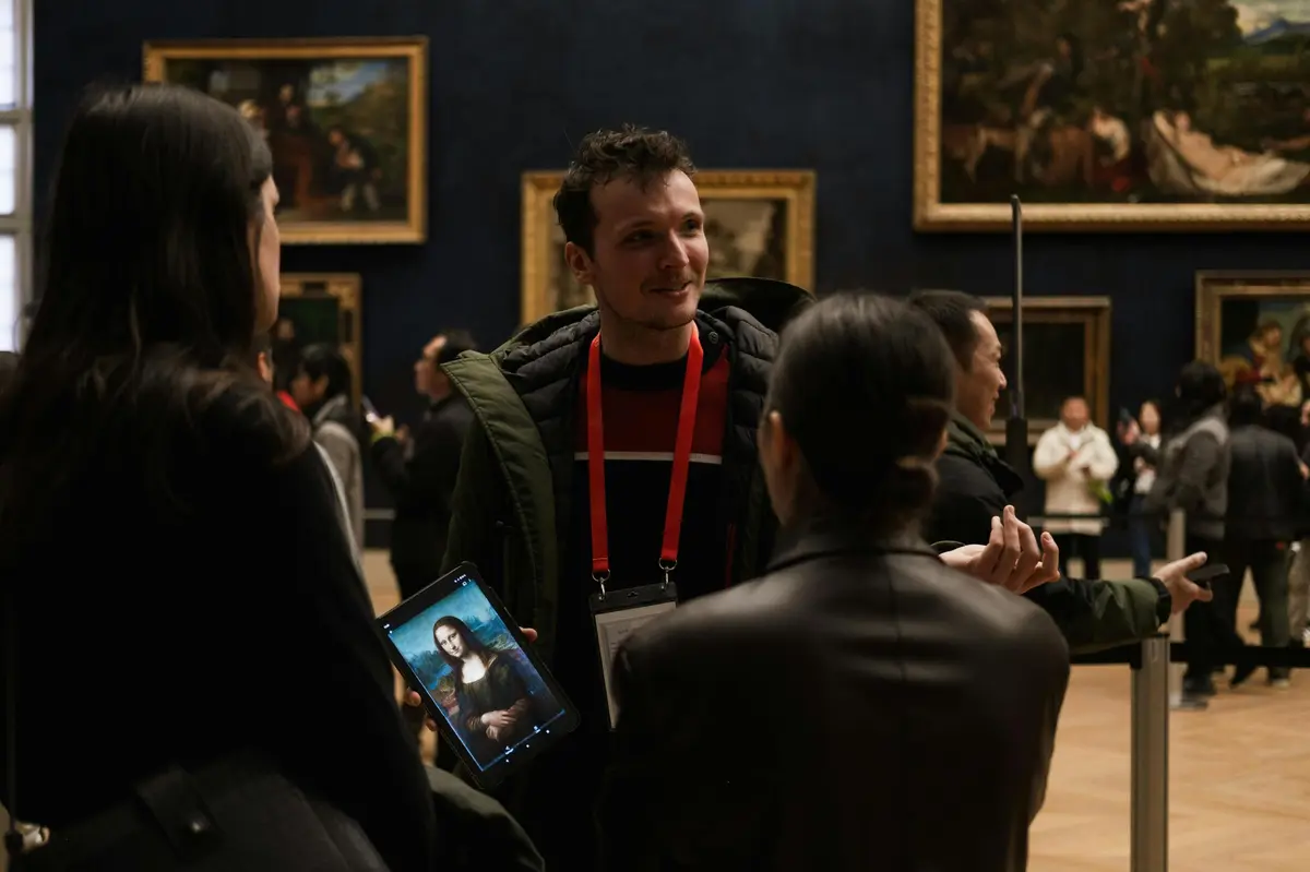 Un homme tenant une tablette affichant la Joconde, discutant avec une femme dans un musée dont les murs sont couverts d'œuvres d'art.