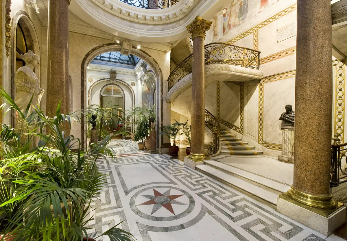 Intérieur élaboré avec sols en marbre, escalier orné, colonnes, plantes en pot et sculptures dans un atrium bien éclairé.