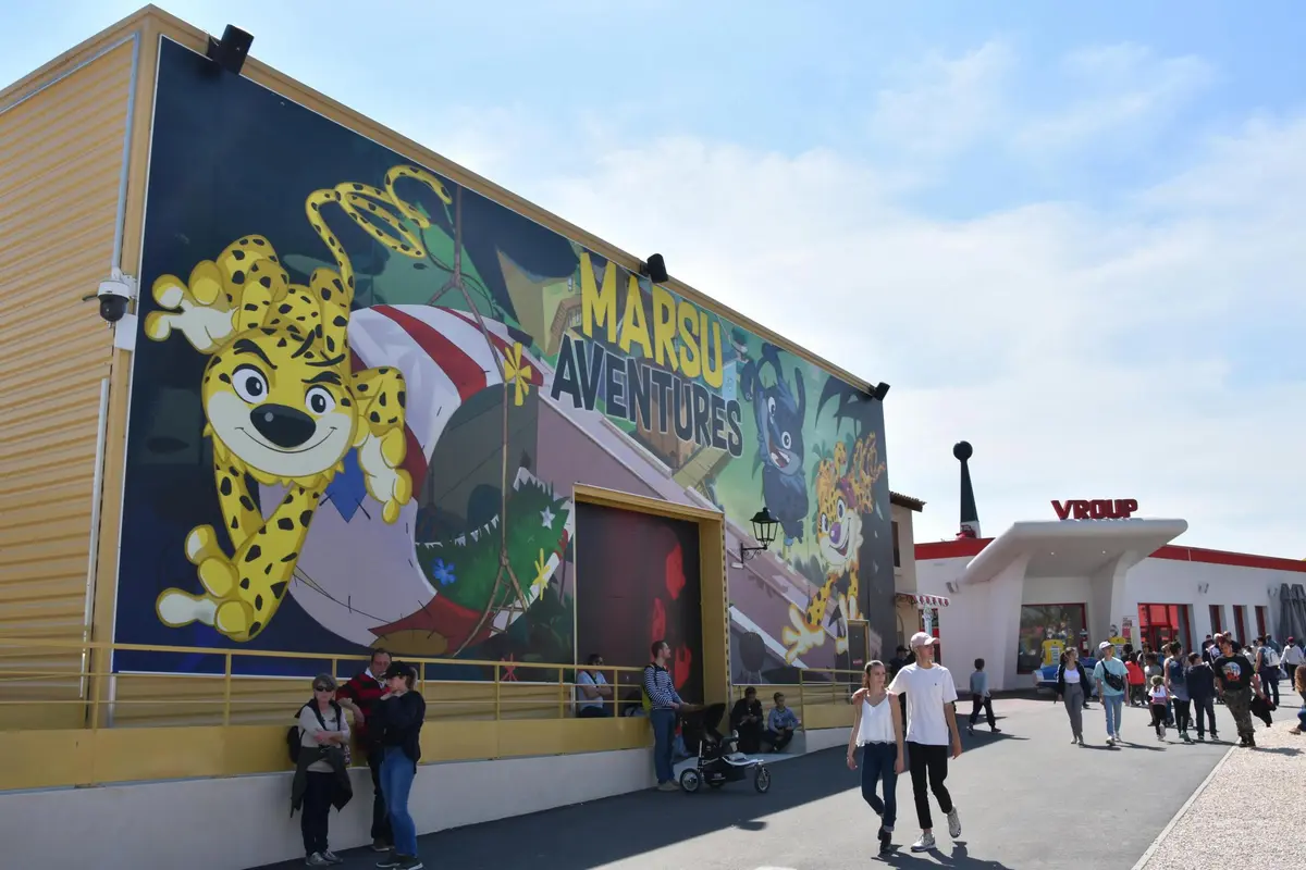 Des personnes marchant près d'un bâtiment avec une grande peinture murale Marsu Aventures et un panneau d'entrée Vroupe lors d'une journée ensoleillée.