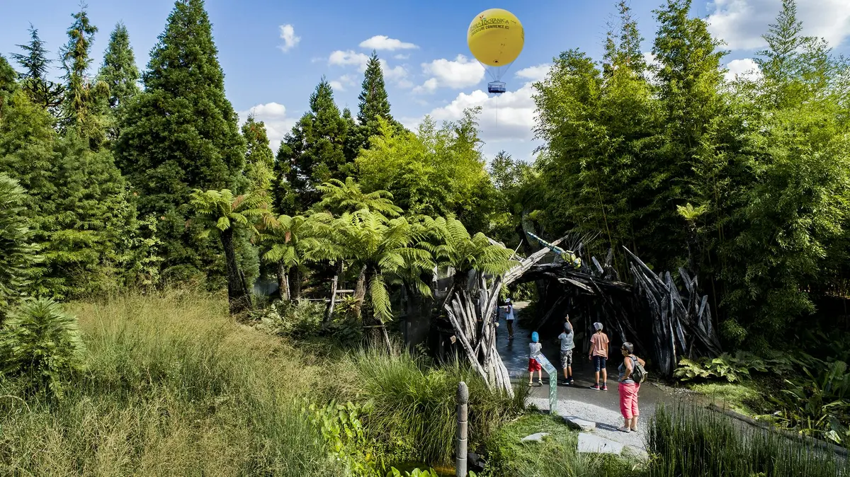 Des enfants et des adultes explorent un parc verdoyant avec une arche de branches en bois ; une montgolfière jaune flotte au-dessus de leur tête.