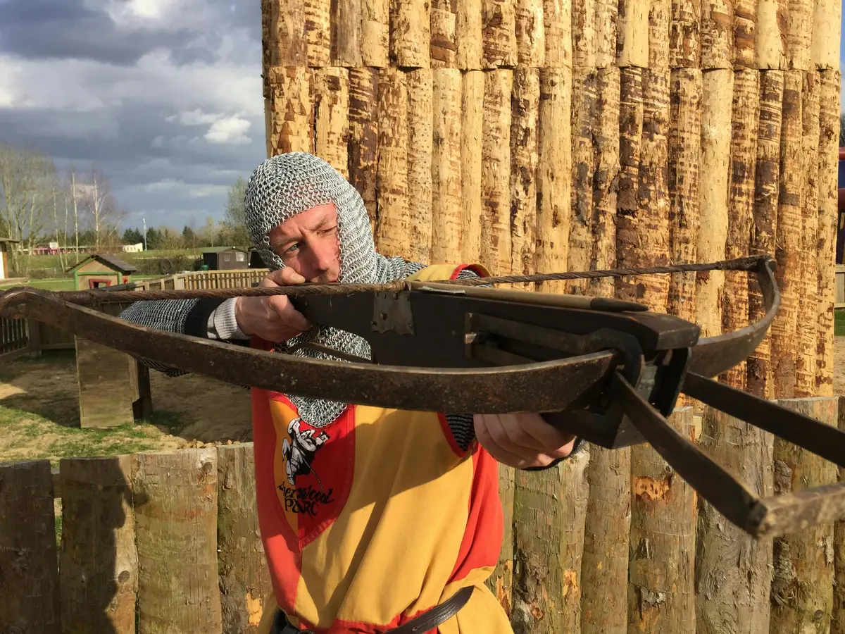 Une personne en cotte de mailles et en tenue médiévale visant une grande arbalète sur un site de reconstitution historique en plein air.