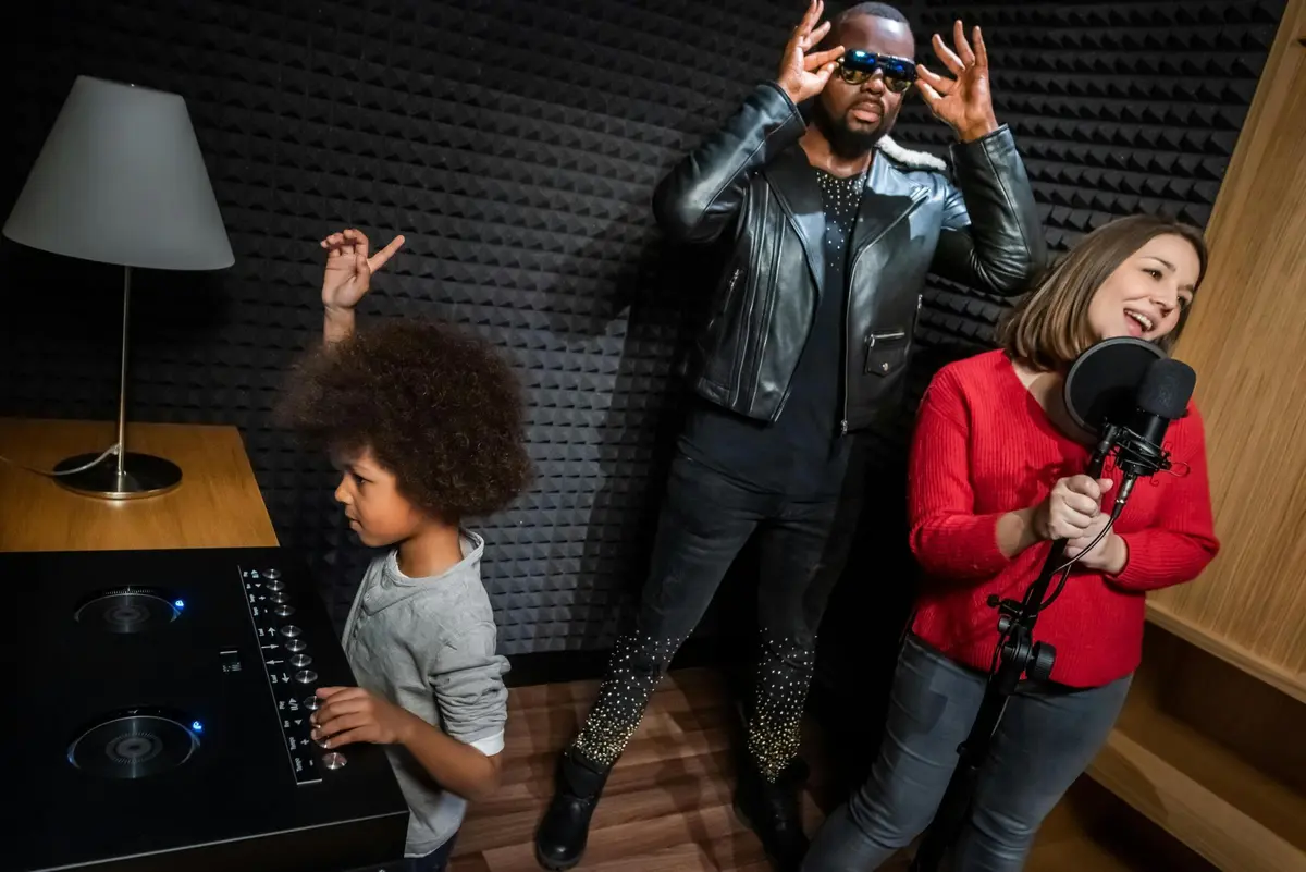 Trois personnes dans un studio d'enregistrement : un enfant qui joue, un homme avec des lunettes de soleil qui ajuste ses lunettes et une femme qui chante dans un microphone.