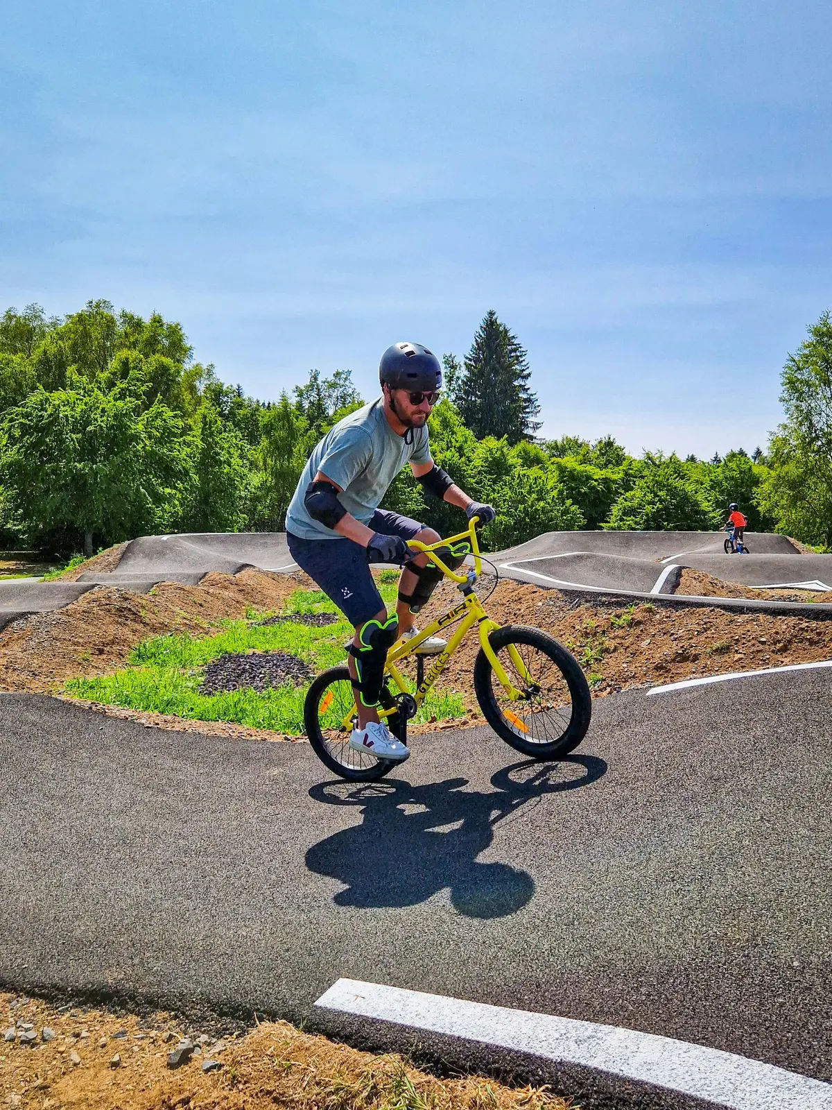 Une personne utilise un vélo BMX jaune sur une piste de pompage dans un parc ensoleillé, en portant un casque et un équipement de protection.