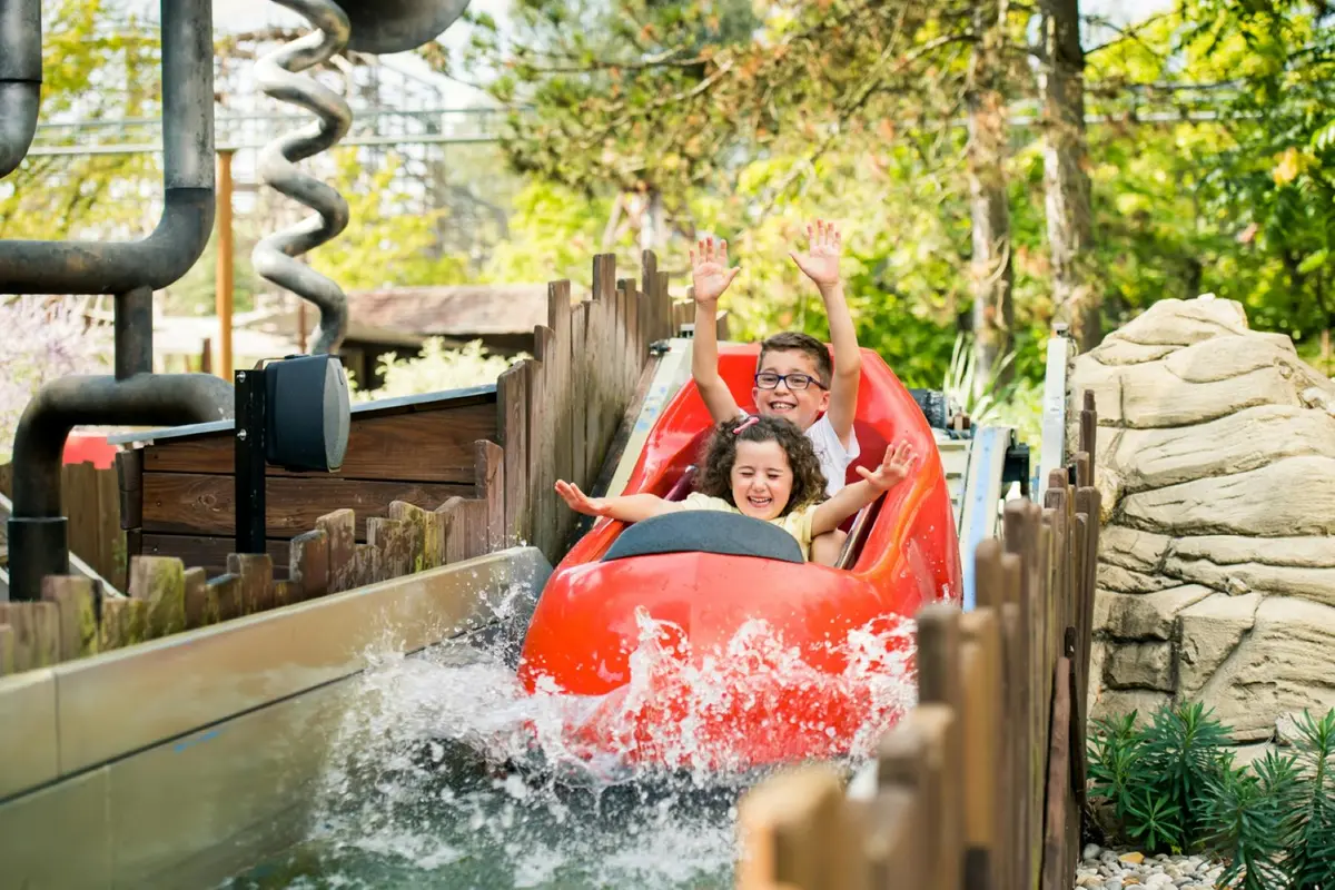 Deux enfants souriants sur un toboggan de bois rouge, les bras levés, alors qu'ils s'éclaboussent dans l'eau. Verdure environnante et clôture en bois.