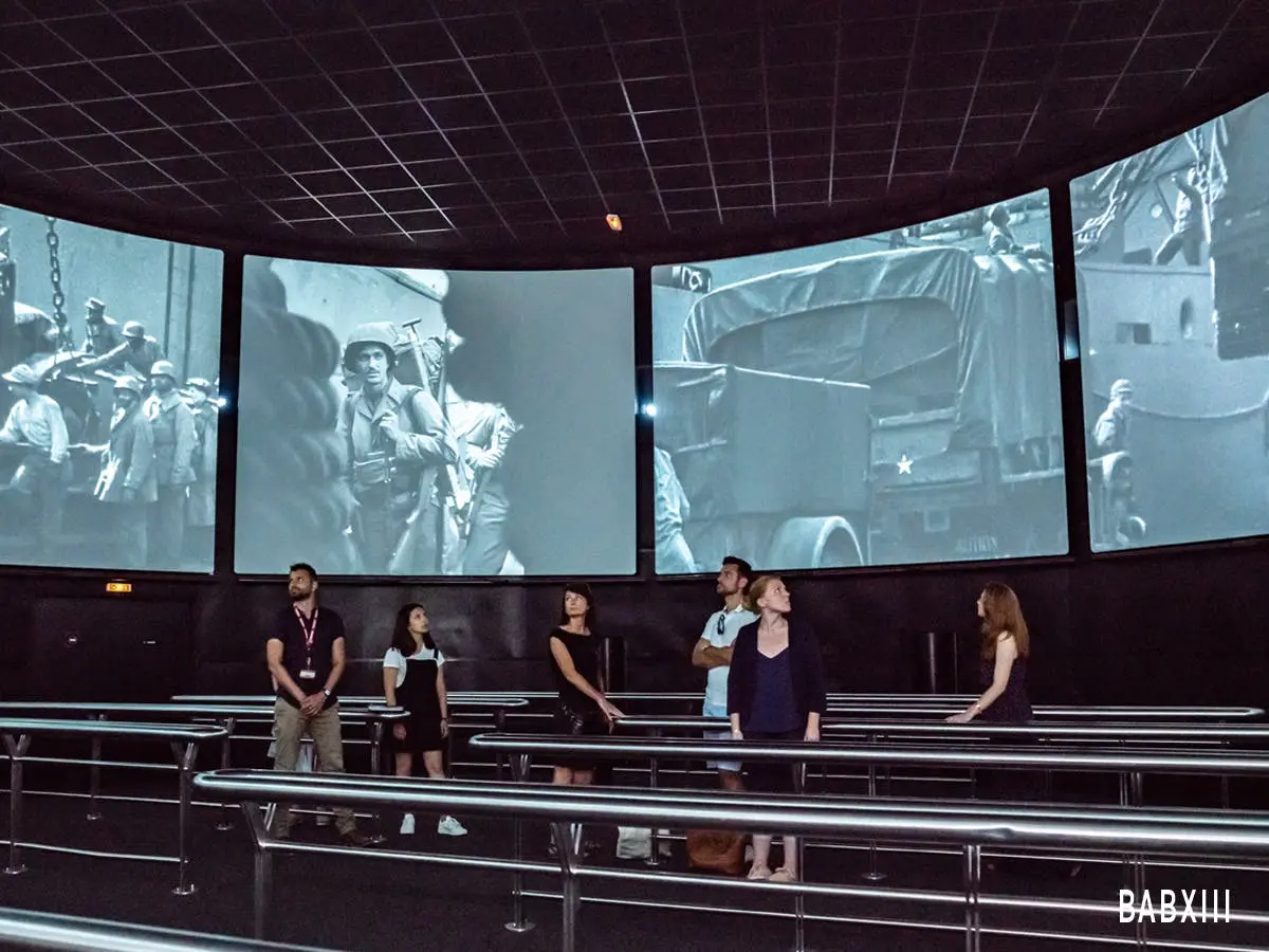 Des personnes dans une pièce sombre avec de grands écrans muraux diffusant des images de guerre en noir et blanc.
