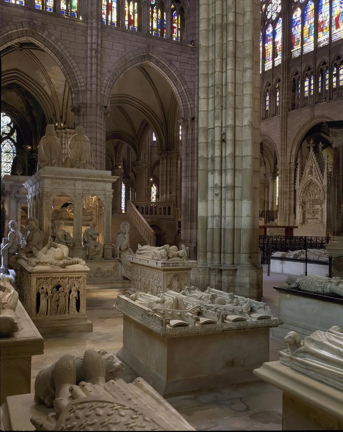 Un intérieur de cathédrale avec de nombreux tombeaux en pierre, des statues et des éléments architecturaux ornés sous des plafonds voûtés.