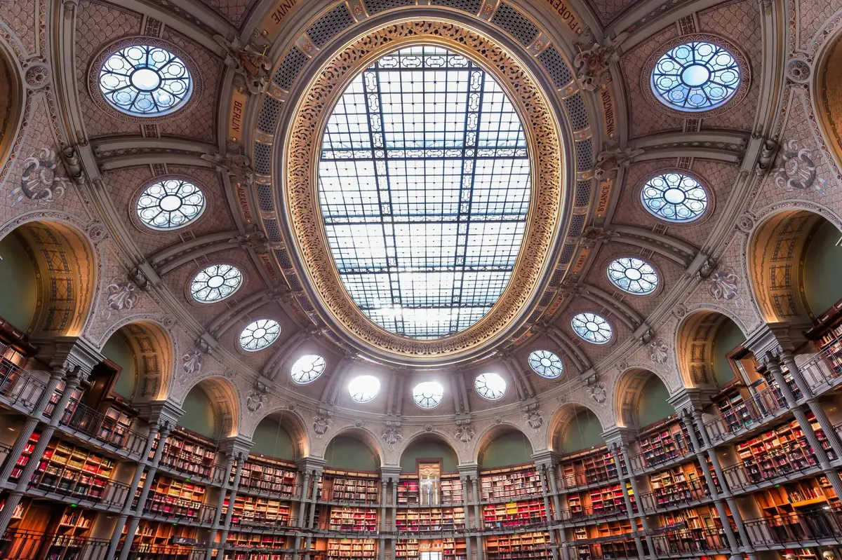 L'intérieur d'une grande bibliothèque avec de hautes étagères et un grand plafond ovale en verre teinté, entouré de décorations ornementales.
