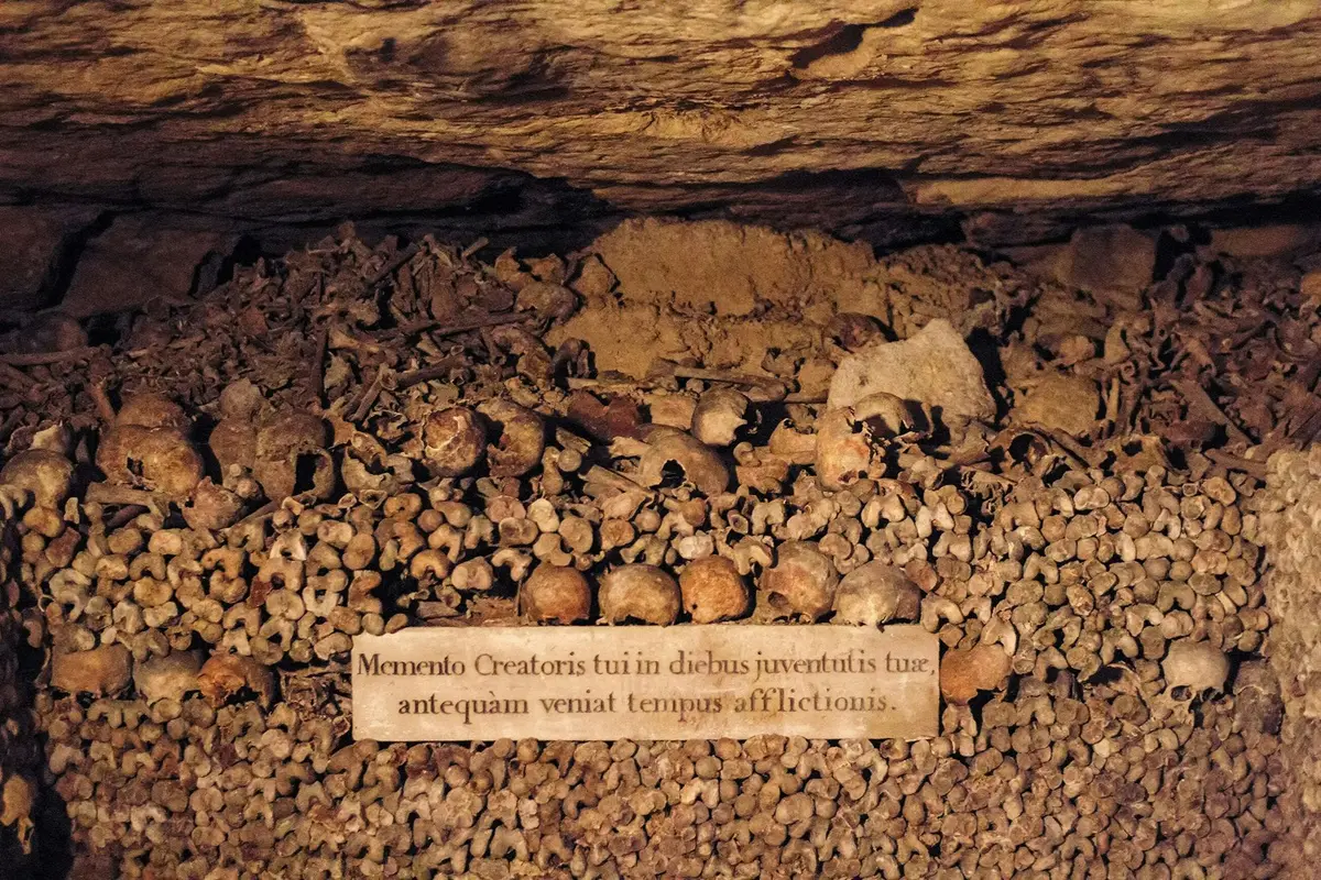 Un tas d'os et de crânes humains disposés dans une catacombe souterraine, avec une inscription en latin sur une plaque.