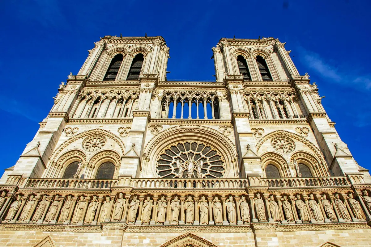 La façade de la cathédrale Notre-Dame avec ses tours jumelles, ses sculptures ornées, son grand vitrail circulaire et ses statues sur fond de ciel bleu.