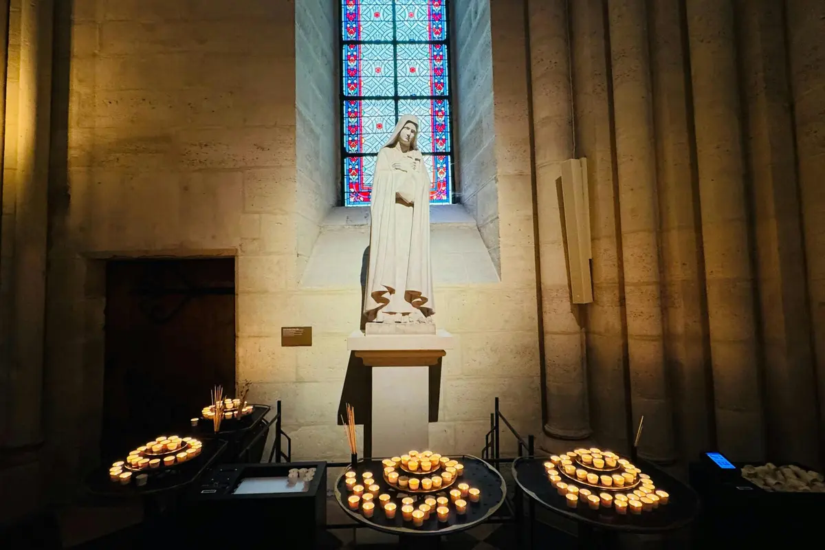 Une statue d'un personnage vêtu se tient devant un vitrail, entouré de bougies allumées sur des supports circulaires.