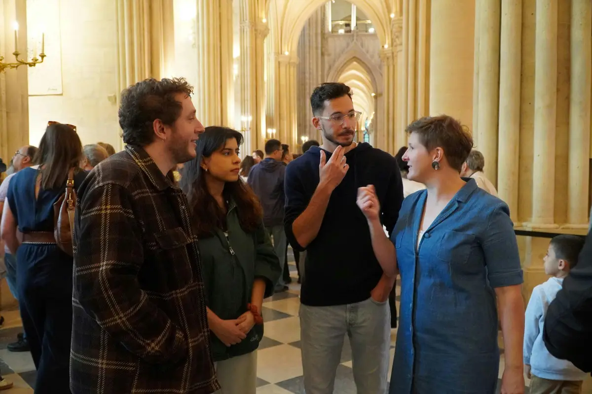Groupe de personnes discutant à l'intérieur d'un grand couloir voûté avec de hautes colonnes et un sol en damier.