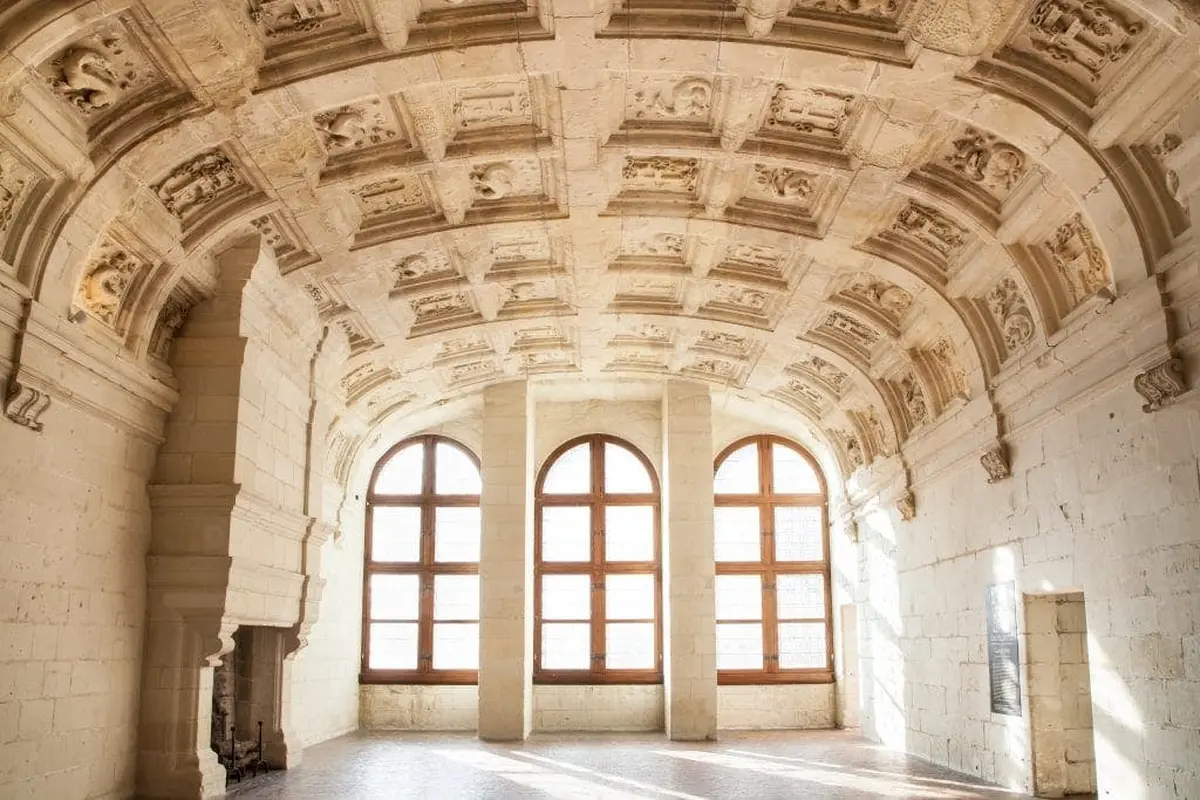 Un plafond voûté orné de sculptures complexes, trois grandes fenêtres cintrées et la lumière du soleil qui pénètre dans une pièce vide au sol en pierre.