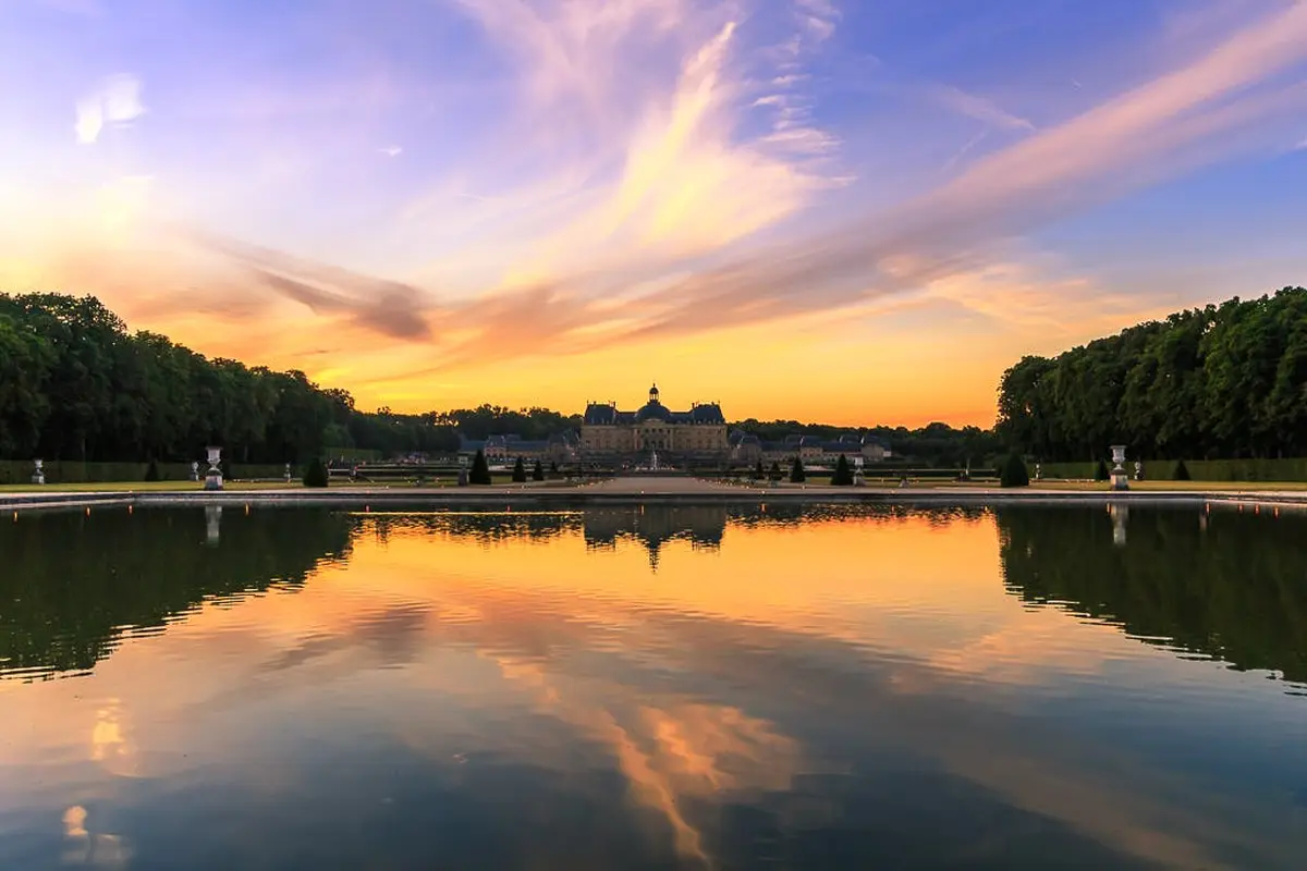Un grand bâtiment au coucher du soleil, se reflétant dans un étang calme avec des nuages vibrants et des silhouettes d'arbres environnants.
