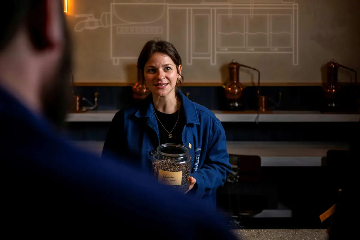 Une femme en veste bleue tient une jarre et parle, avec un bar et un décor en laiton en arrière-plan.