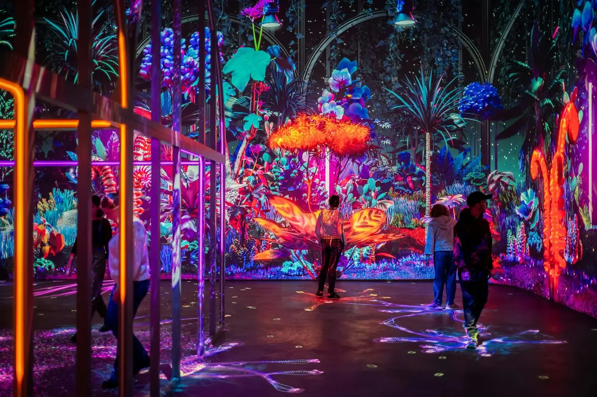 Des personnes se promènent dans une installation artistique intérieure colorée et illuminée, composée de plantes et de projections lumineuses.