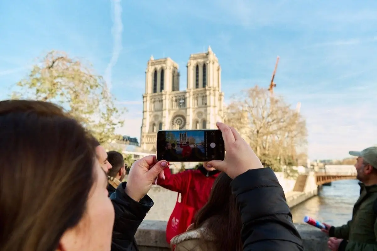 Une personne brandit un smartphone pour prendre une photo de la cathédrale Notre-Dame, avec d'autres personnes et une grue visibles à l'arrière-plan.