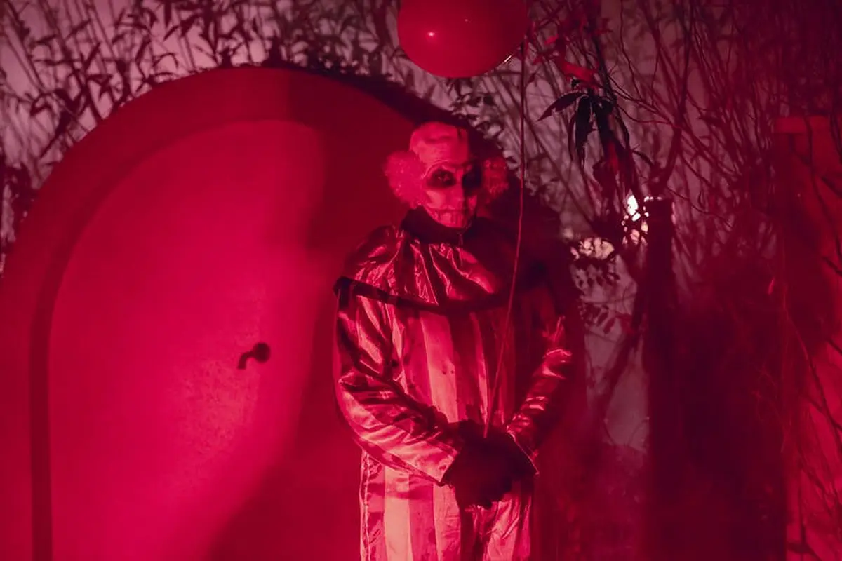 Un clown à l'allure sinistre, éclairé par une lumière rouge, tenant un ballon au milieu d'un feuillage sombre et d'ombres.