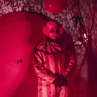 Un clown à l'allure sinistre, éclairé par une lumière rouge, tenant un ballon au milieu d'un feuillage sombre et d'ombres. DR