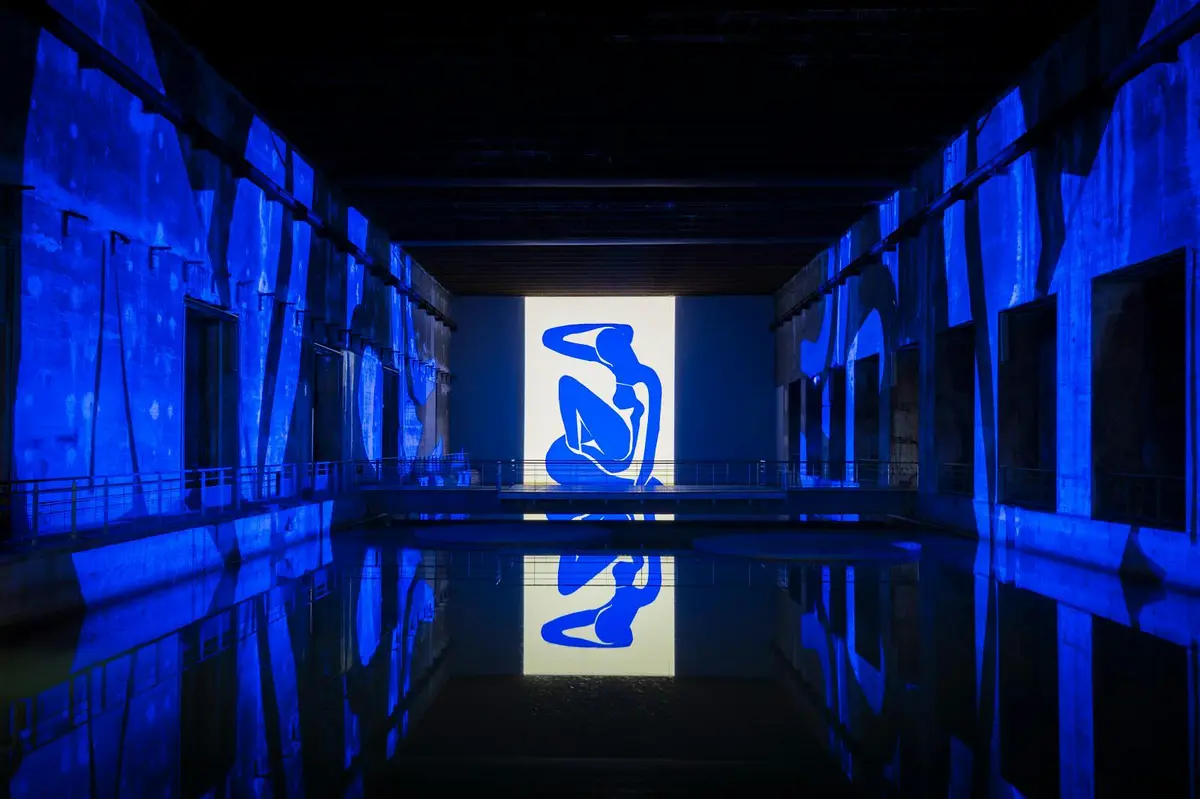 Une figure abstraite bleue projetée sur un mur dans un espace faiblement éclairé, avec de l'eau réfléchissante en dessous et des environs illuminés.