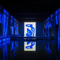 Une figure abstraite bleue projetée sur un mur dans un espace faiblement éclairé, avec de l'eau réfléchissante en dessous et des environs illuminés. &copy; @Culturespaces / Geoffroy Groult