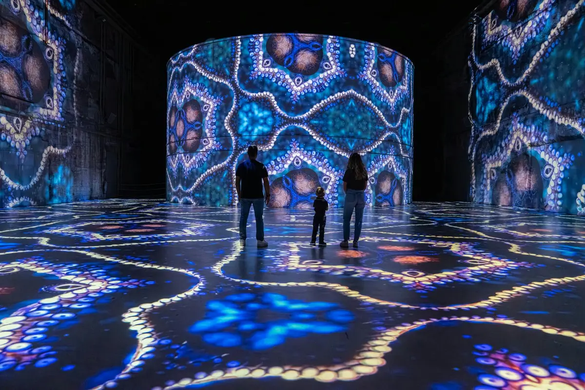 Trois personnes se trouvent dans une pièce sombre éclairée par de grandes projections lumineuses colorées et kaléidoscopiques sur le sol et les murs.