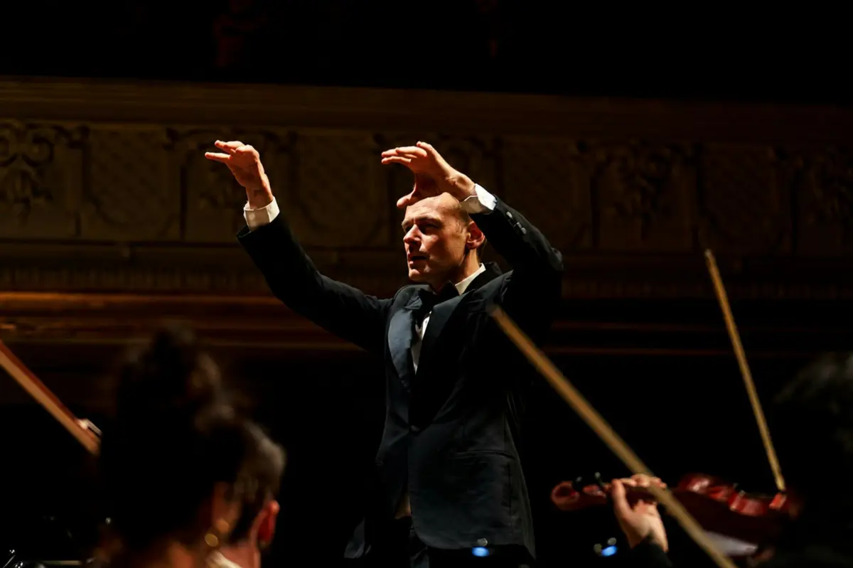 Un chef d'orchestre en tenue de cérémonie dirigeant un concert, avec des musiciens et des archets visibles au premier plan.