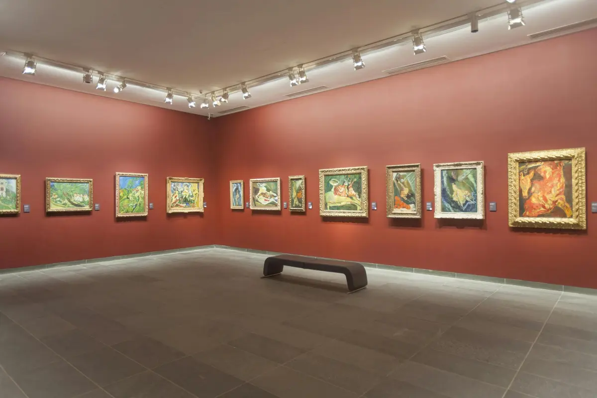 Galerie de musée aux murs rouges où sont exposés des tableaux encadrés. Un seul banc est posé sur le sol carrelé au centre de la pièce.