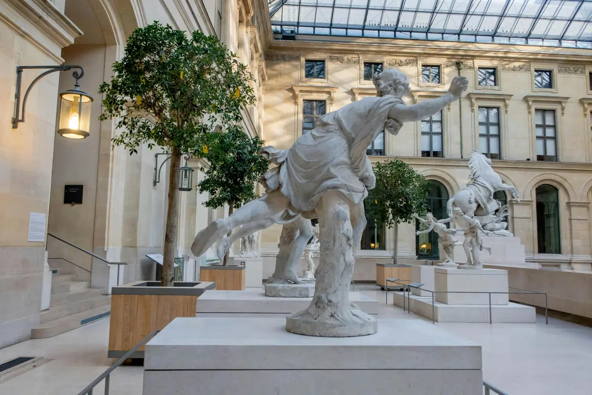 Une statue en marbre d'un personnage dynamique dans un atrium de musée avec des arbres et un plafond en verre.