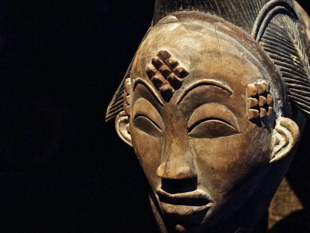 Masque en bois sculpté avec des motifs géométriques sur le front et les joues, sur fond sombre.