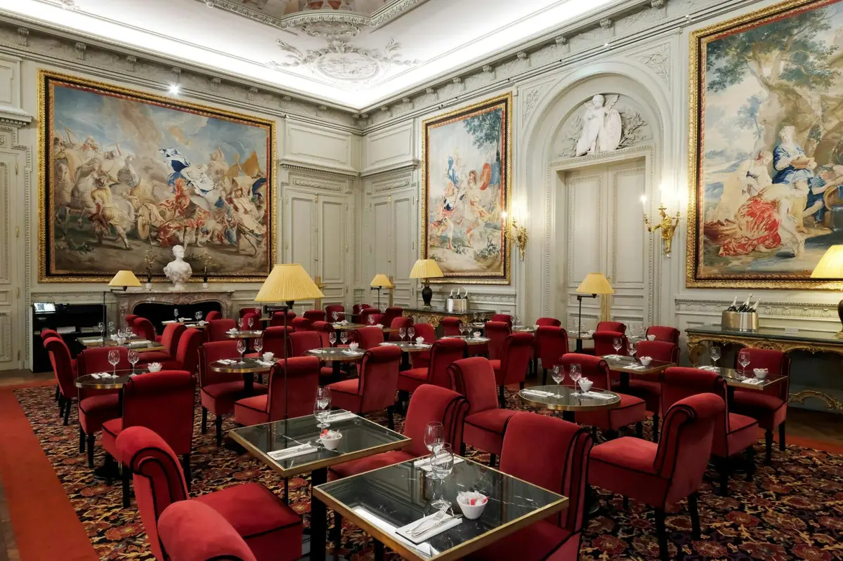 Salle à manger élégante avec des chaises rembourrées rouges, des tables à plateau en verre, des œuvres d'art murales, un plafond orné et un éclairage doux provenant de lampes et d'appliques.