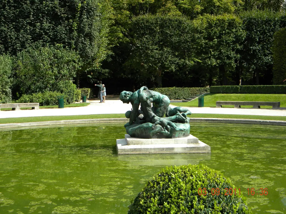 Statue en bronze représentant des personnages entrelacés sur un piédestal au centre d'un étang vert, entouré d'arbres et d'arbustes dans un parc.