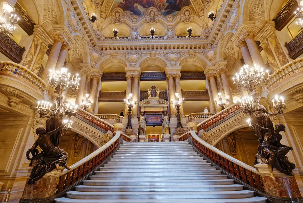 Grand escalier avec des rampes ornées, des lustres et des colonnes dans un hall opulent, richement décoré et doté de hauts plafonds.