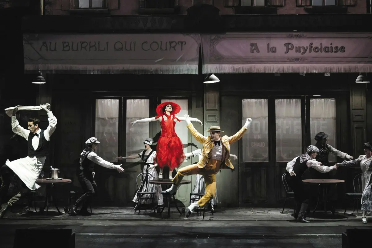 Des artistes aux costumes éclatants dansent sur une scène ressemblant à un café parisien, avec des vitrines détaillées en arrière-plan.