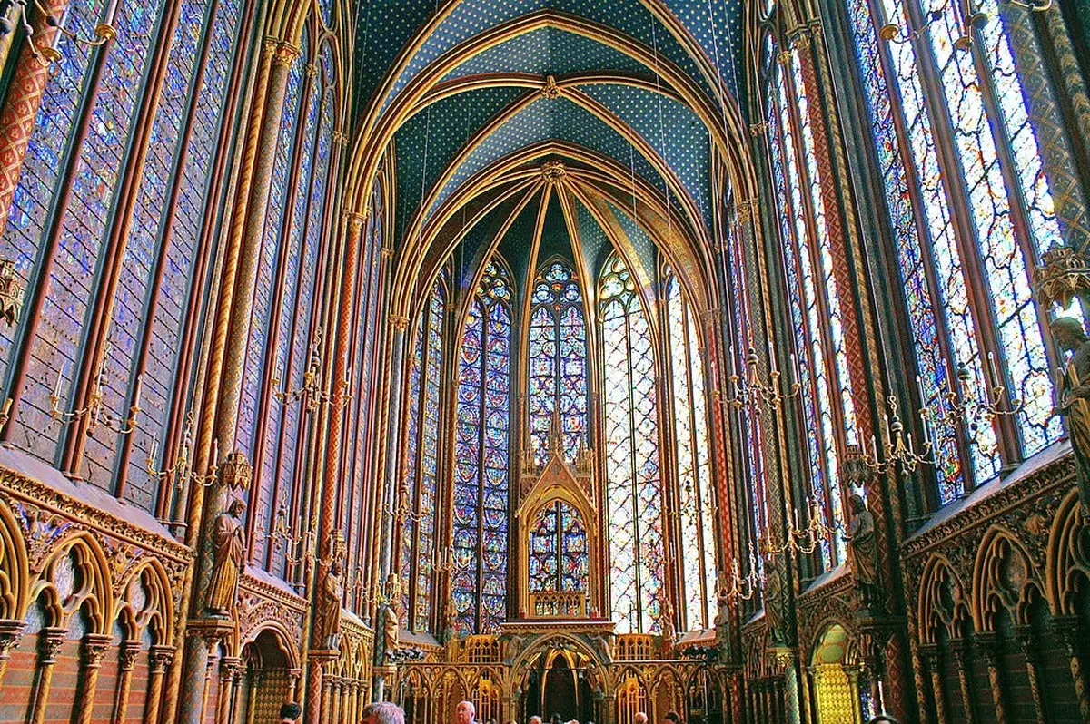 Intérieur de cathédrale orné de hauts vitraux, de plafonds voûtés, de lustres et de statues. Personnes en bas.