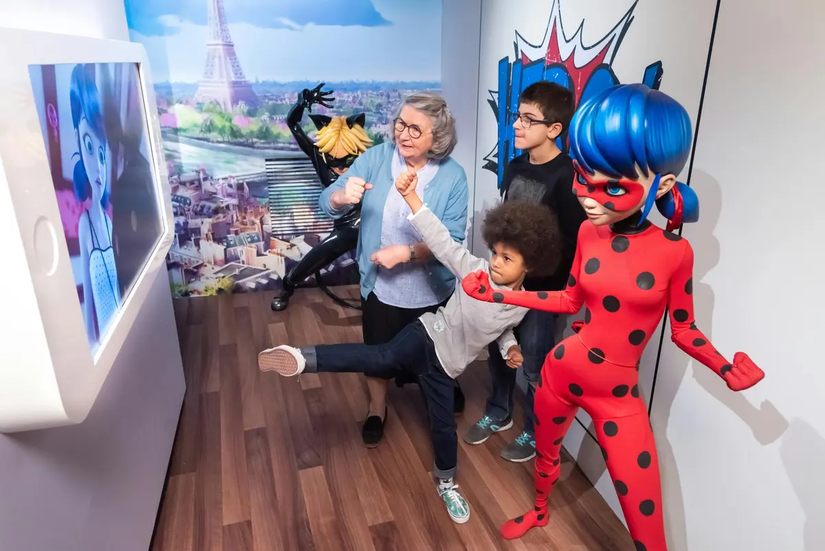Une femme, un garçon et un enfant posent avec les personnages costumés Ladybug et Cat Noir, tous prenant des poses ludiques de super-héros à l'intérieur.