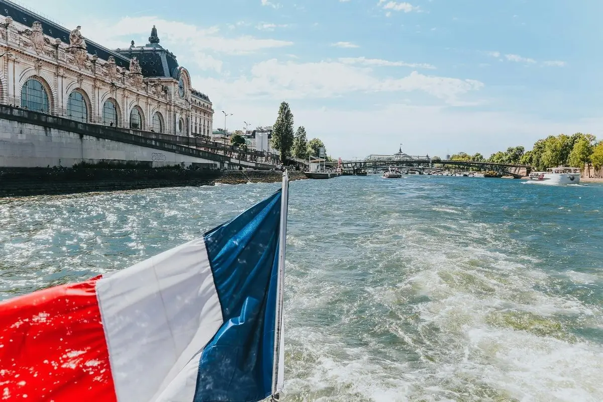 Un drapeau français flotte à la poupe d'un bateau sur une rivière, avec en arrière-plan un bâtiment historique et un pont.