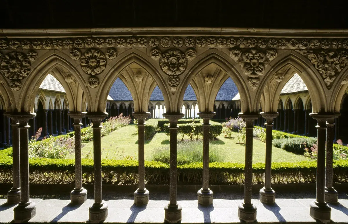 Des arches de pierre ornées encadrent une cour de jardin avec des buissons et des fleurs soignés sous un ciel ensoleillé.