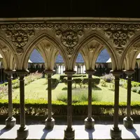 Des arches de pierre ornées encadrent une cour de jardin avec des buissons et des fleurs soignés sous un ciel ensoleillé. DR