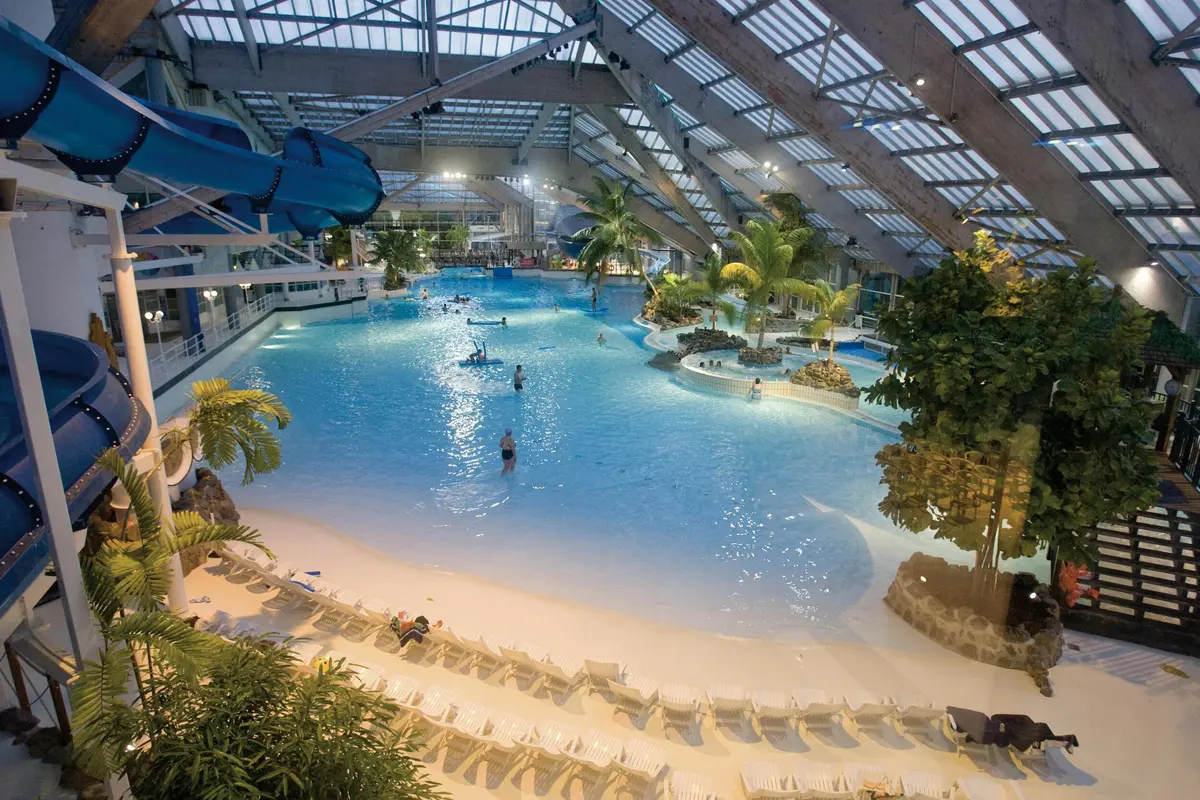 Parc aquatique intérieur avec une grande piscine, des palmiers, des chaises longues et un toboggan bleu. Plusieurs personnes sont dans l'eau.