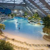 Parc aquatique intérieur avec une grande piscine, des palmiers, des chaises longues et un toboggan bleu. Plusieurs personnes sont dans l'eau. DR