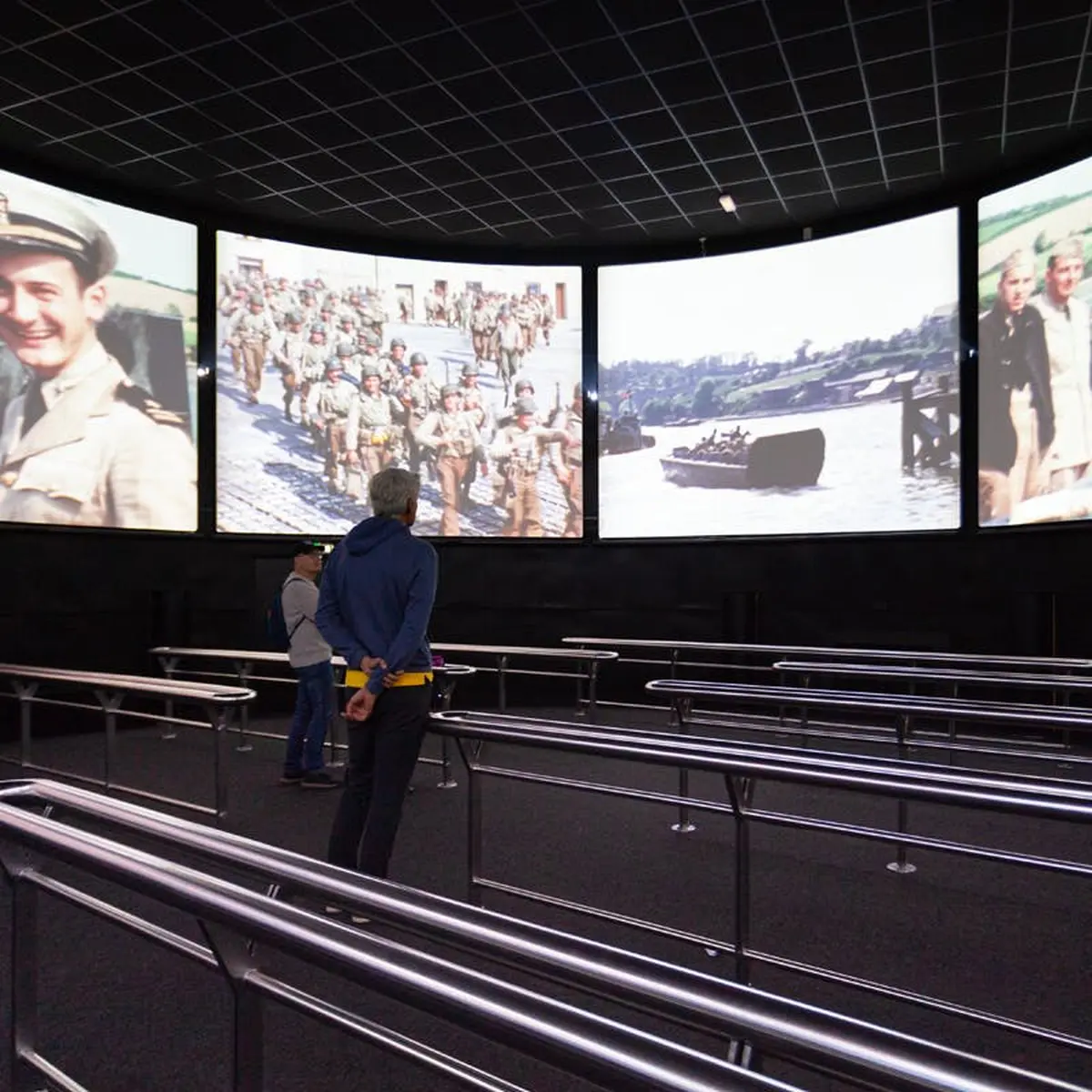 Deux personnes debout devant quatre grands écrans diffusant des séquences militaires historiques dans une salle obscure avec des grilles métalliques.