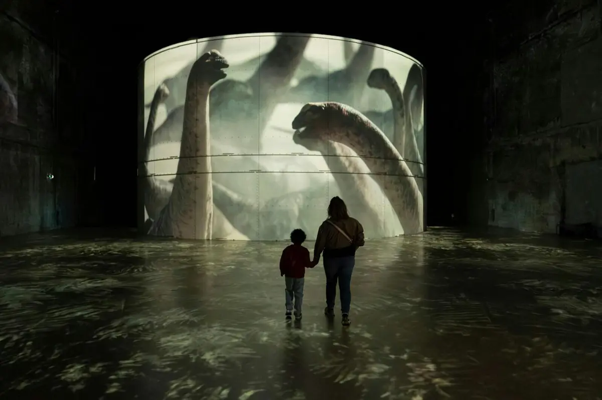 Une personne et un enfant se tenant par la main, face à un grand écran circulaire sur lequel sont projetés des dinosaures, dans une salle d'exposition sombre.