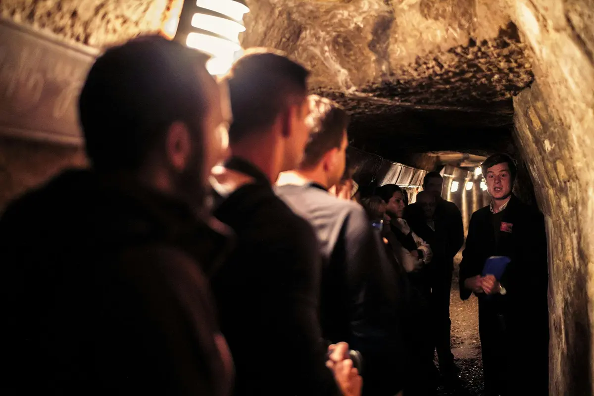 Un groupe de personnes se trouvant dans un tunnel souterrain étroit et faiblement éclairé, avec un guide qui leur parle en tenant des notes à la main.