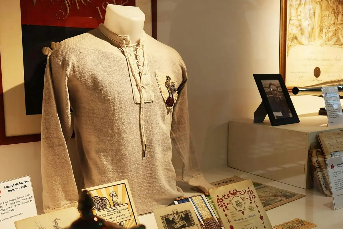 Vitrine de musée présentant une chemise beige à dentelle ornée d'un emblème, des documents d'époque et, en arrière-plan, une photo encadrée.