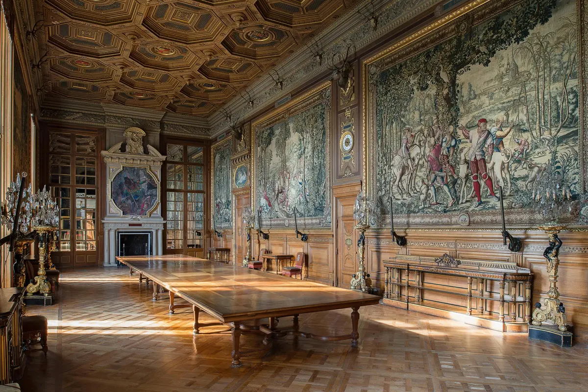 Salle ornée d'une longue table en bois, de tapisseries complexes sur les murs, d'un plafond détaillé et d'une cheminée.