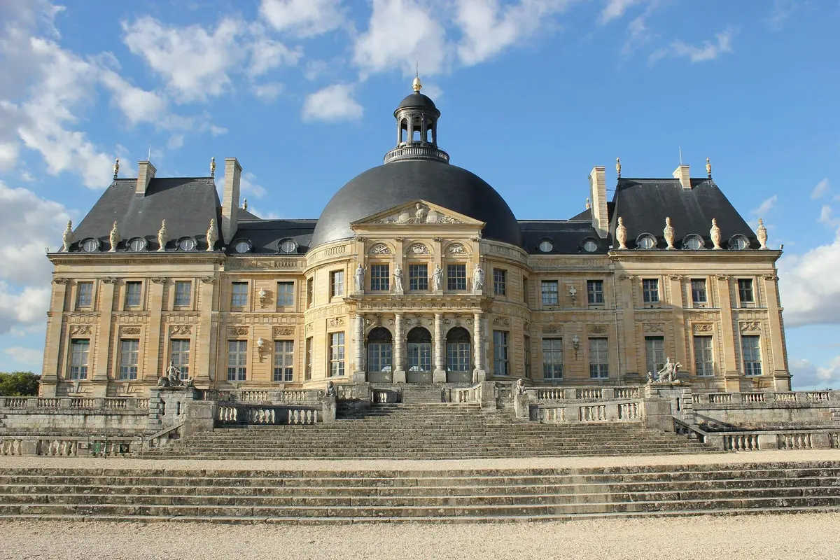 A grand French château with a large dome, ornate architecture, and stone steps leading to the entrance under a blue sky.