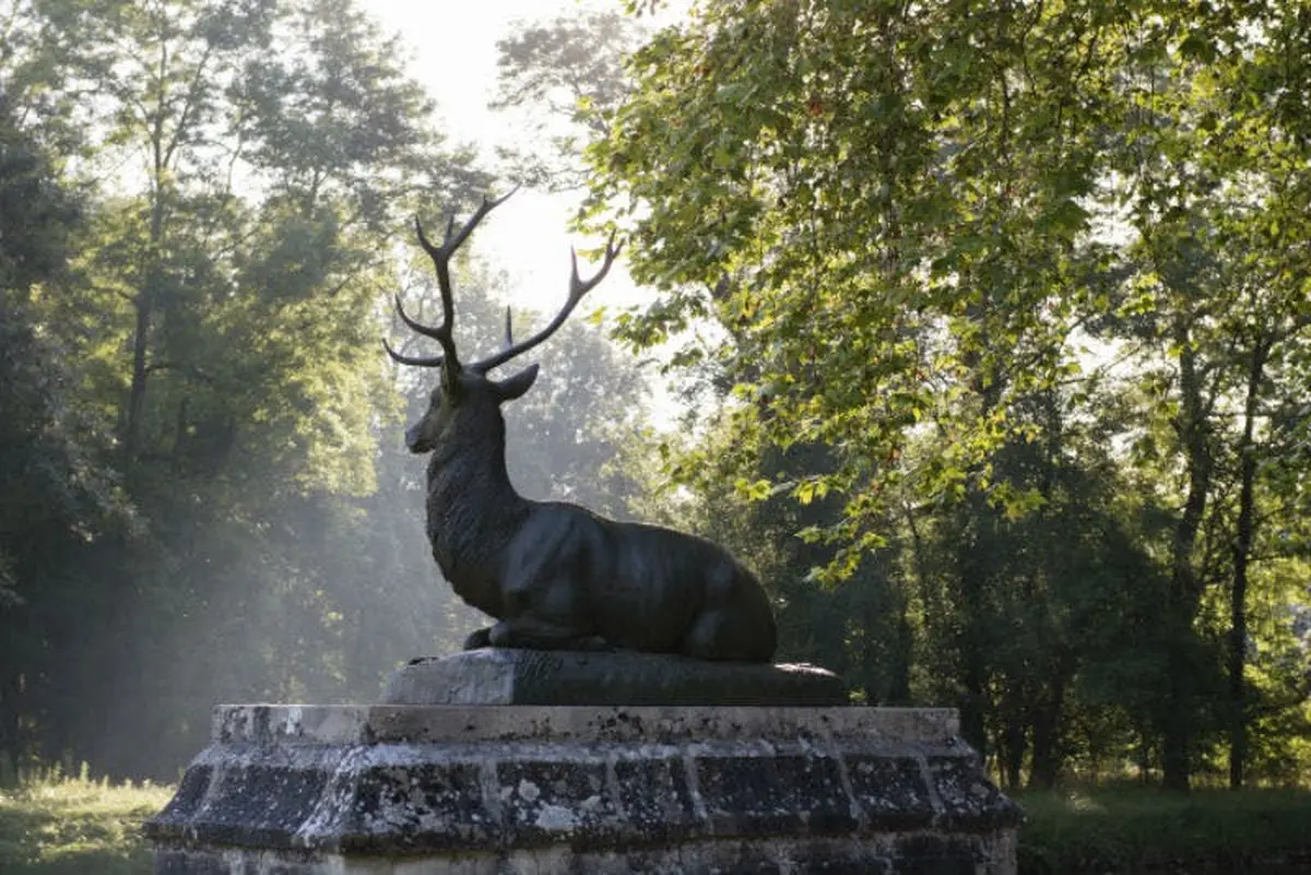 Statue d'un cerf couché avec des bois sur un piédestal en pierre, entouré d'arbres dans un parc ensoleillé.