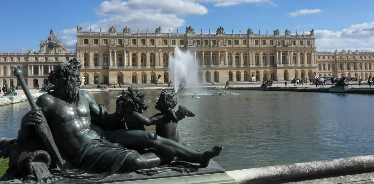Statues in front of a large fountain and the Palace of Versailles under a partly cloudy sky.