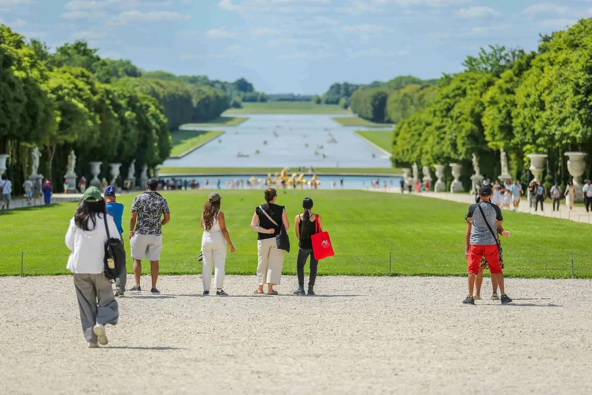 Des personnes se dirigent vers un grand bassin rectangulaire dans un jardin bien entretenu, bordé d'arbres et de statues, sous un ciel bleu.