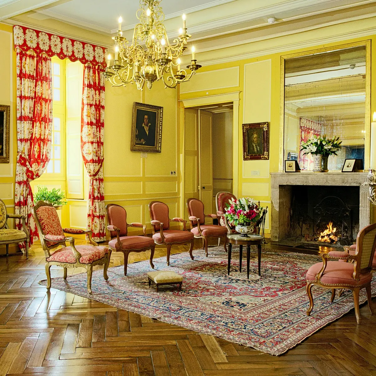 Chambre luxueuse avec des murs jaunes, des meubles ornés, un lustre, une cheminée, des rideaux rouges et blancs et un tapis à fleurs.